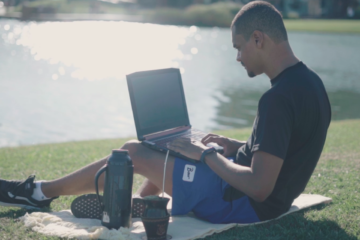 homem sentado na grama com um notebook no colo, ao fundo está um lago
