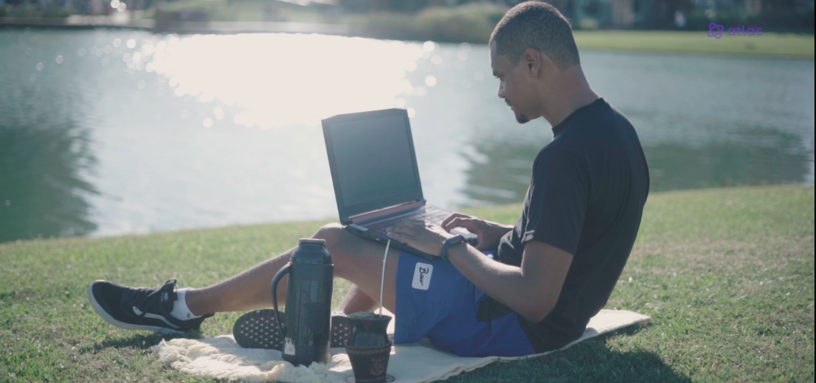 homem sentado na grama com um notebook no colo, ao fundo está um lago